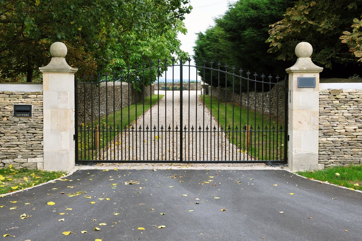 Iron gate with stone pillars opening to a gravel driveway lined with trees and a view of fields beyond