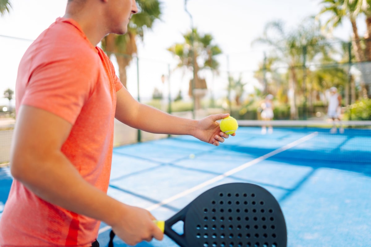 A young man on a blue tennis court holding a racket and ball practicing paddle tennis