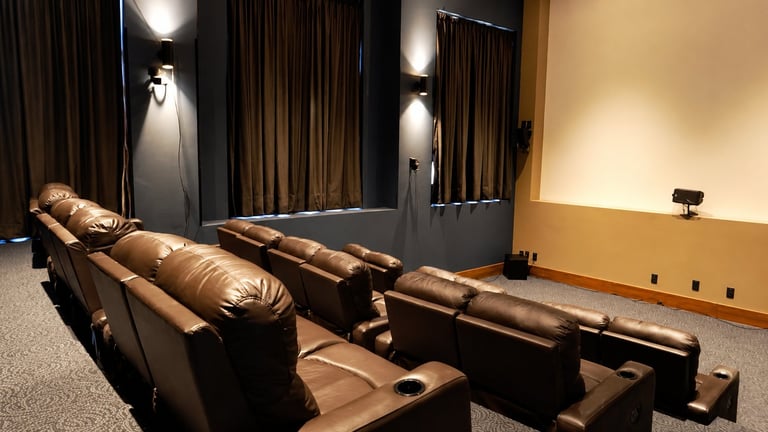 A home theater room with brown leather recliners arranged in rows, warm lighting, and dark curtains on the walls.