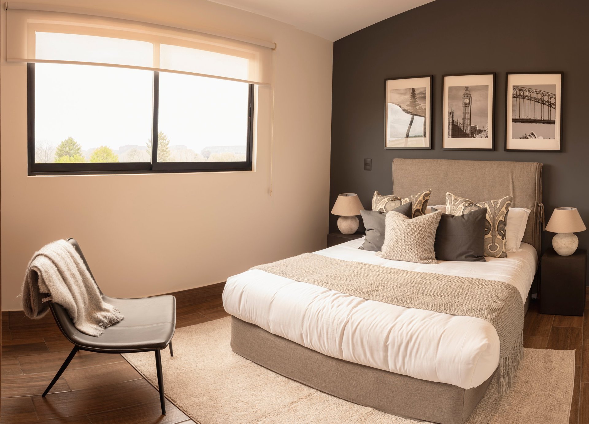 Modern bedroom with beige and brown color scheme, bed with white bedding, accent chair, large window, and framed wall art.
