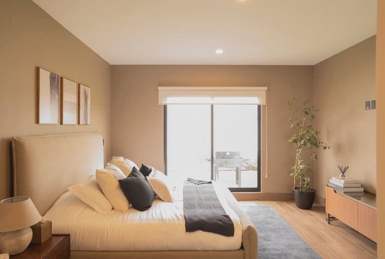 Modern bedroom with beige walls, wooden bed with cream bedding, sliding glass doors, potted plant, and minimalist decor