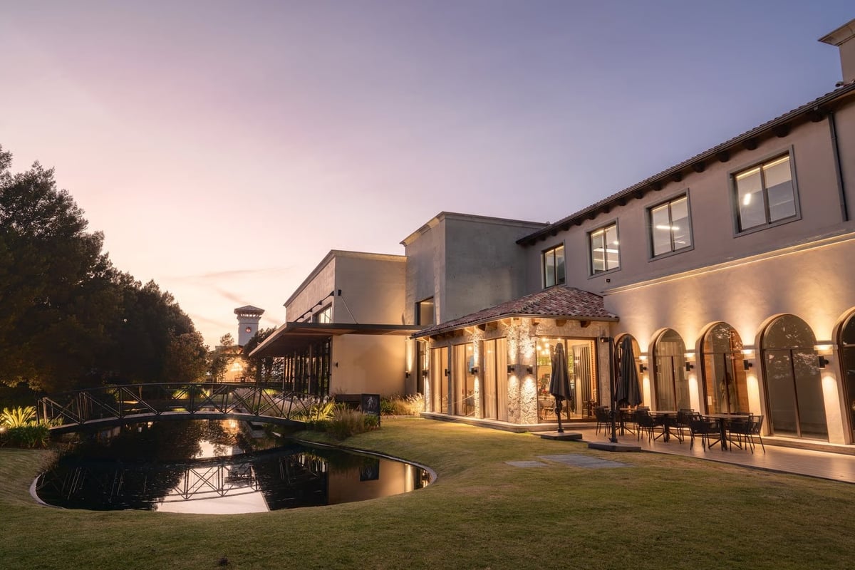 Modern mansion with illuminated arched windows at dusk, reflecting pool in manicured lawn during twilight hour
