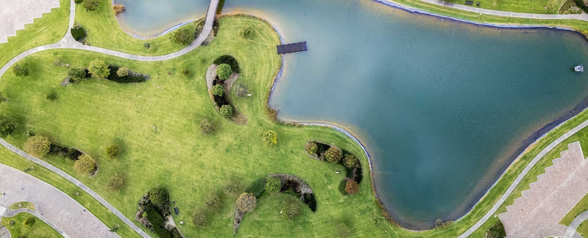 Aerial view of a curved park with green lawn, trees, and a blue pond bordered by paved pathways