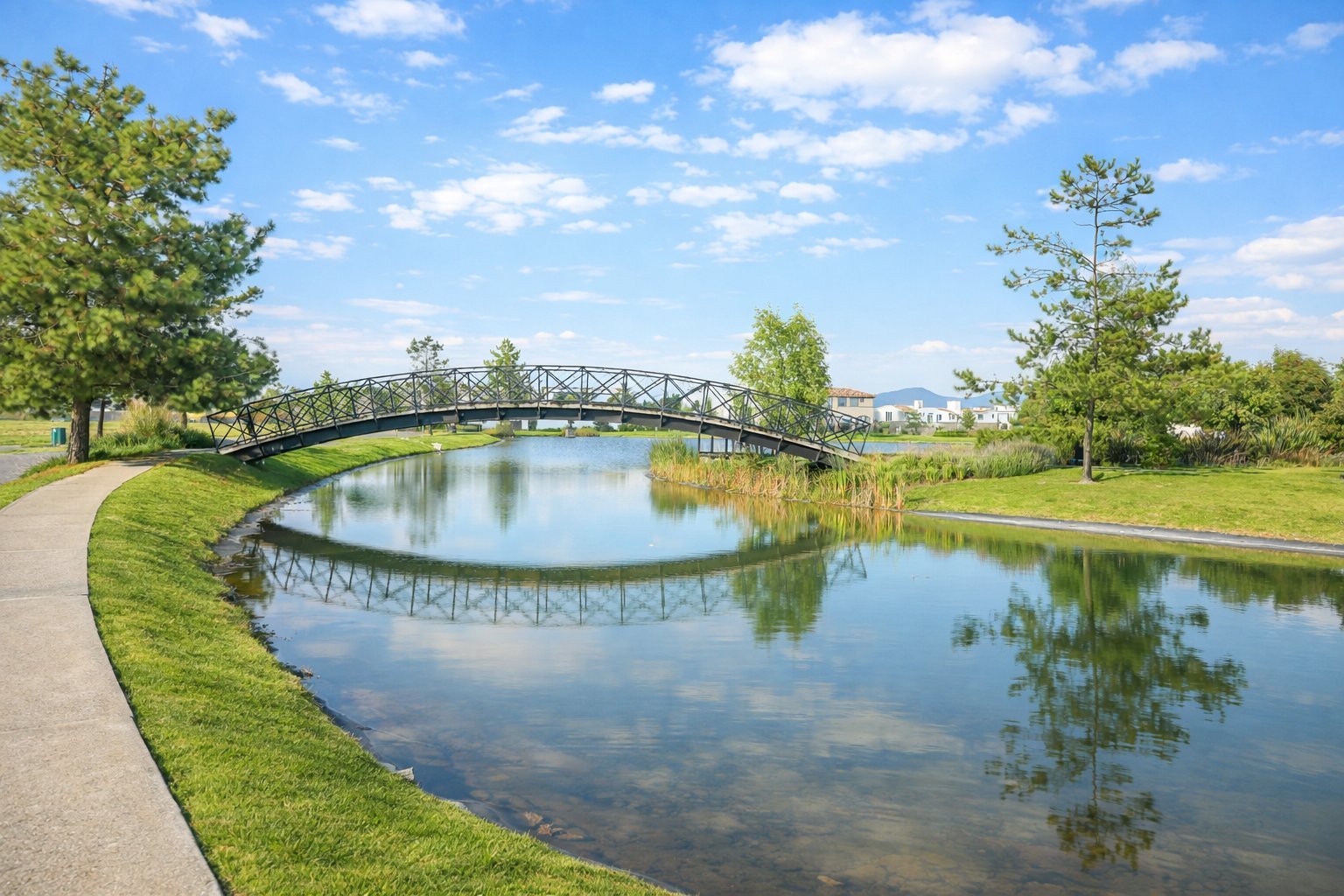Scenic park with metal footbridge spanning calm canal, surrounded by green lawns and trees under clear blue sky