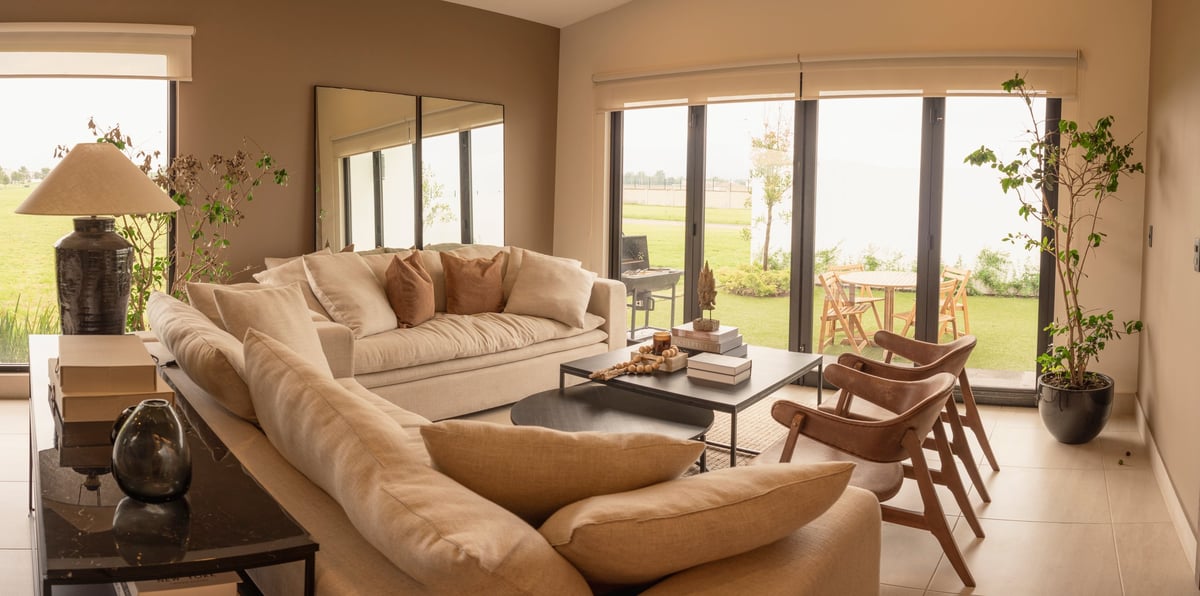 Spacious modern living room with beige sectional sofa, wood furniture, and floor-to-ceiling windows overlooking green landscape.