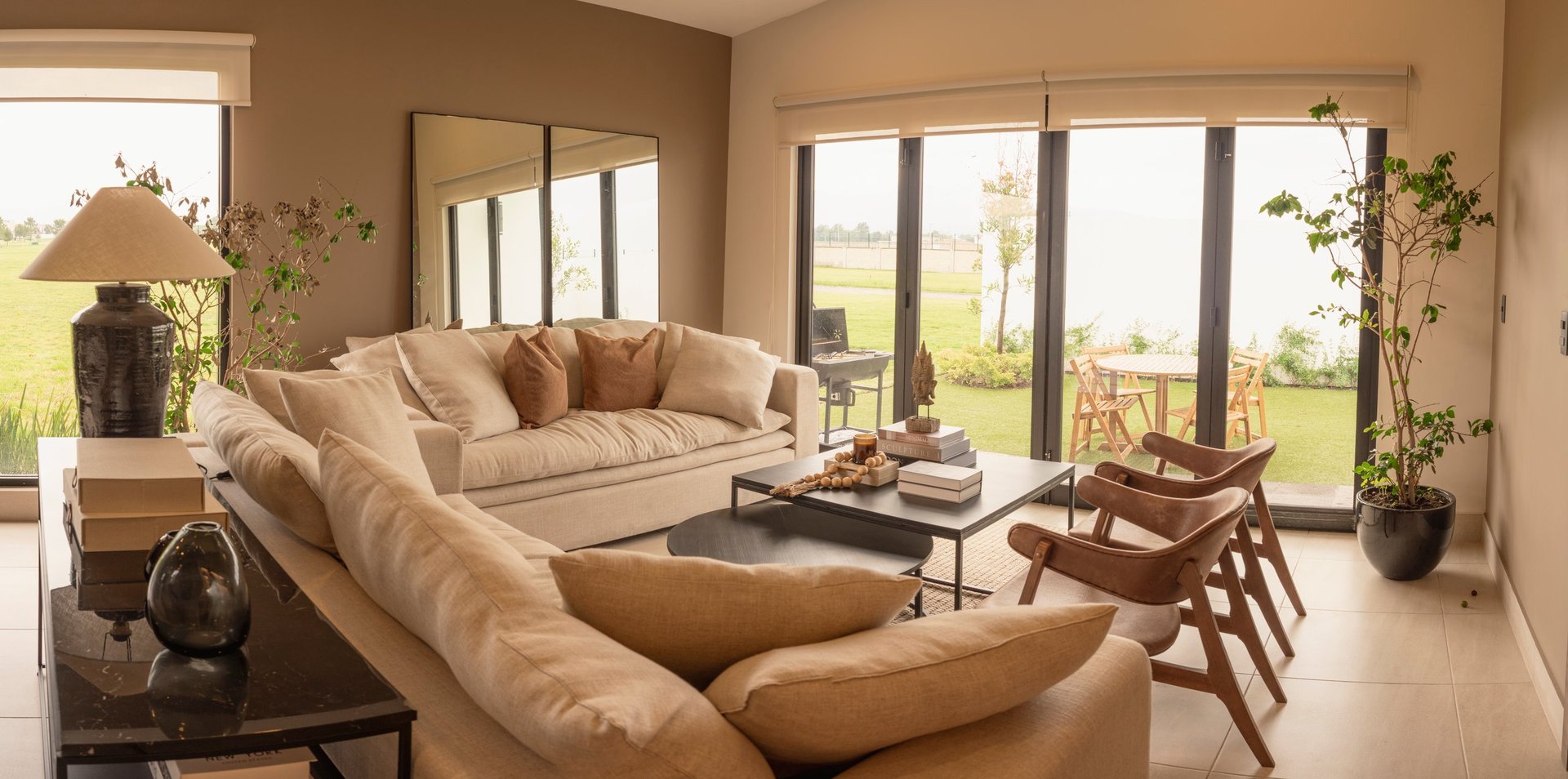 Spacious modern living room with beige sectional sofa, wood furniture, and floor-to-ceiling windows overlooking green landscape.