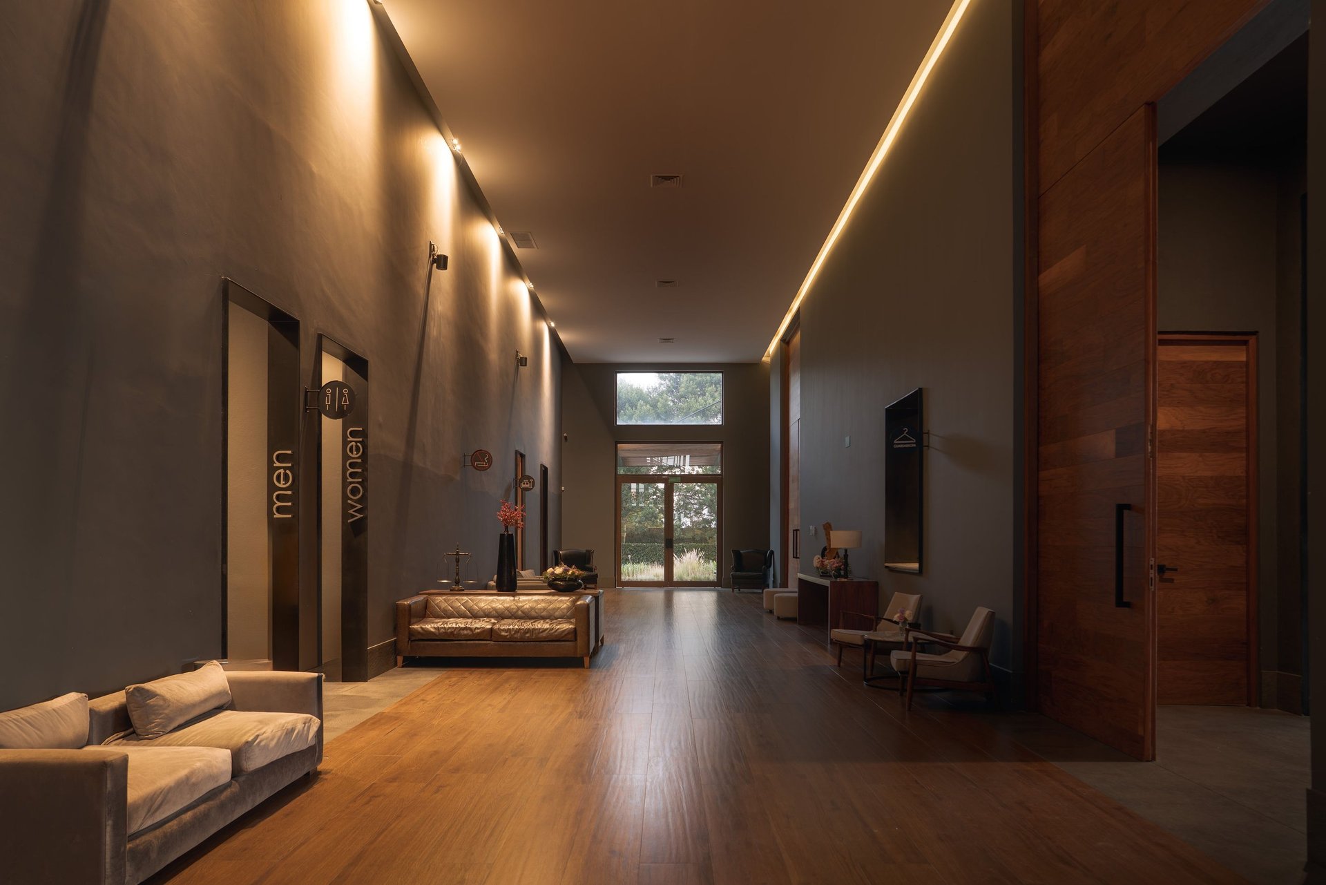 Modern hotel lobby with wooden flooring, linear ceiling lights, warm lighting, elevator doors, seating areas, and natural light from windows