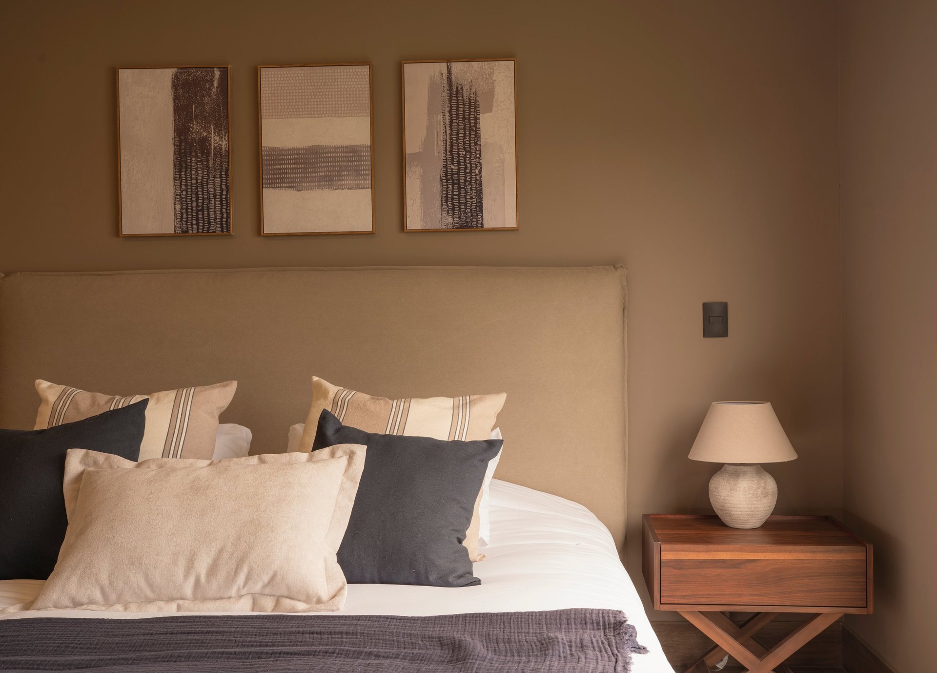 Bedroom with beige walls, bed with cream and dark pillows, wooden nightstand with lamp, and three abstract framed artworks above the headboard