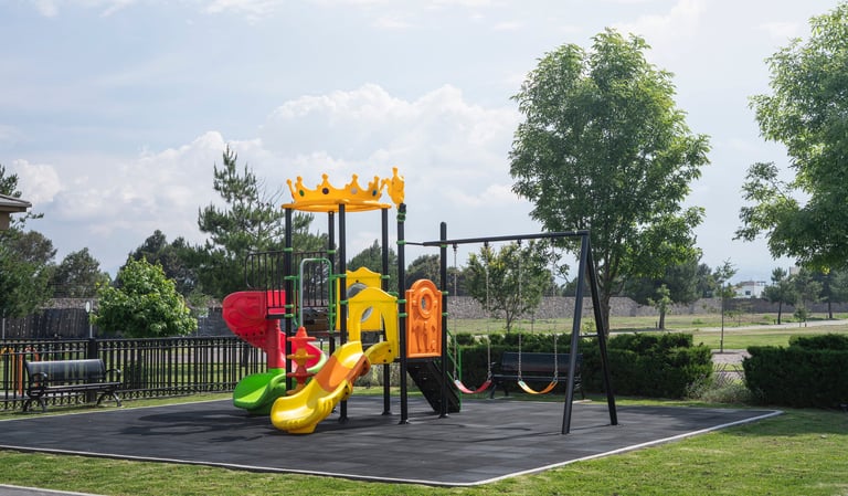 Colorful outdoor playground with yellow, red, and orange equipment including slides and climbing structures on black safety surface, surrounded by grass and trees.