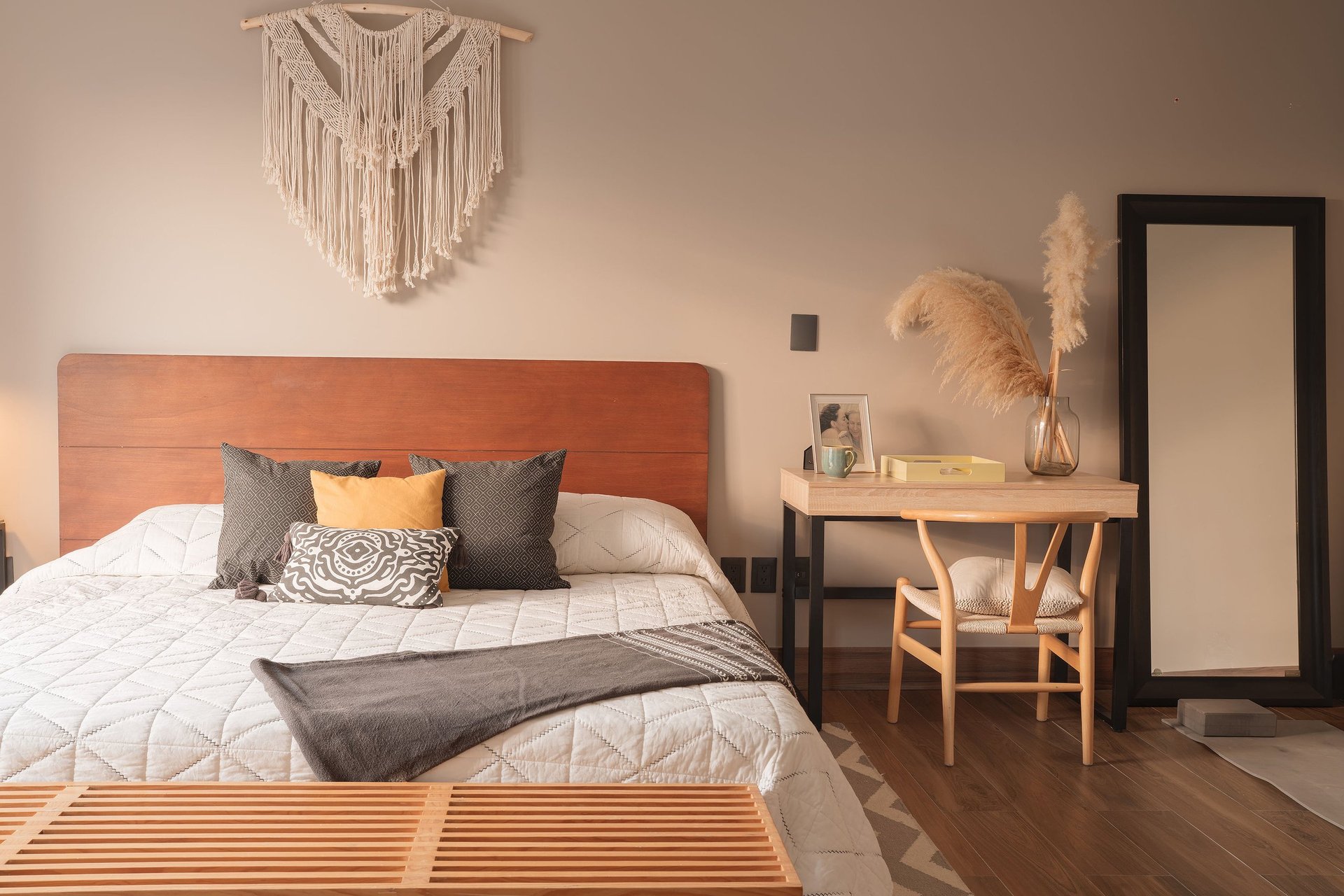 Modern bedroom with terracotta headboard, white bedding, decorative pillows, wooden desk, pampas grass decor, and macramé wall hanging