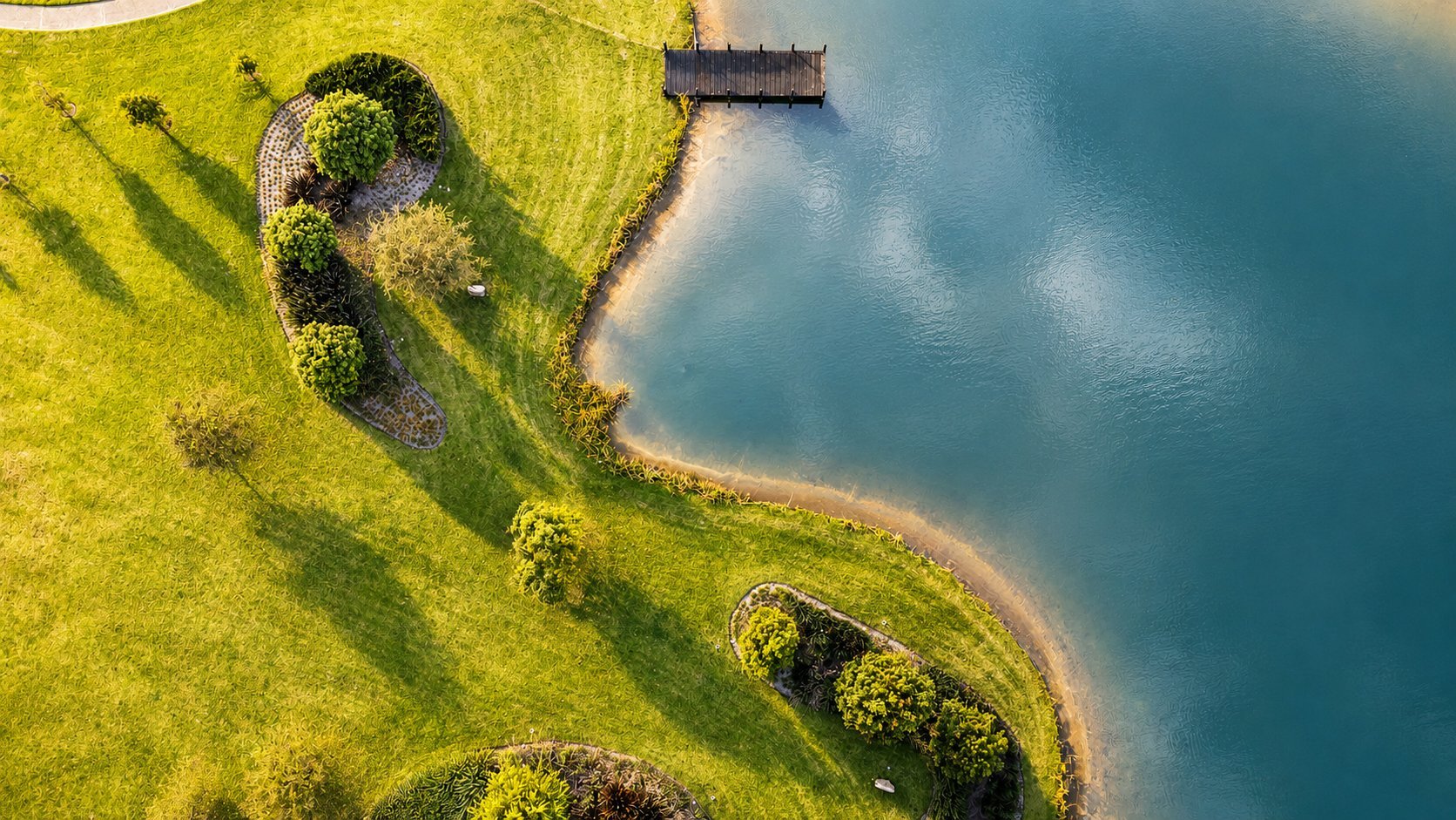 Aerial view of bright green hillside with winding paths meeting turquoise water and a small wooden structure