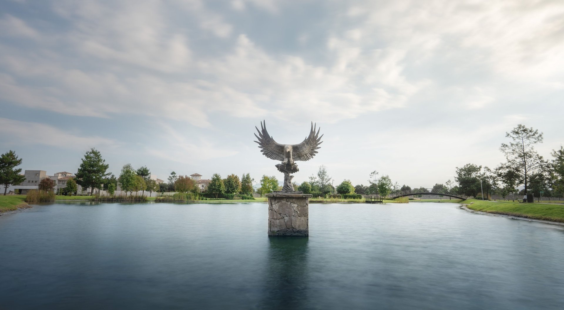 Bronze sculpture of wings spreading upward on a stone pedestal in the center of a calm lake, surrounded by green trees and grassland