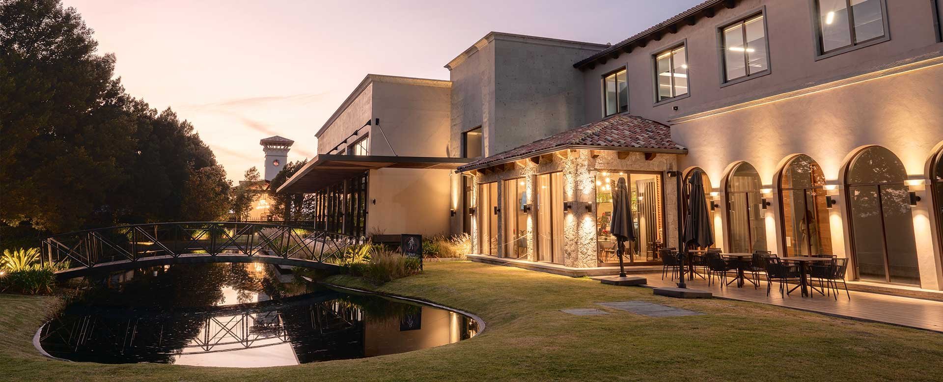 Modern buildings with warm lighting reflected in a pond at dusk, surrounded by manicured landscaping and trees