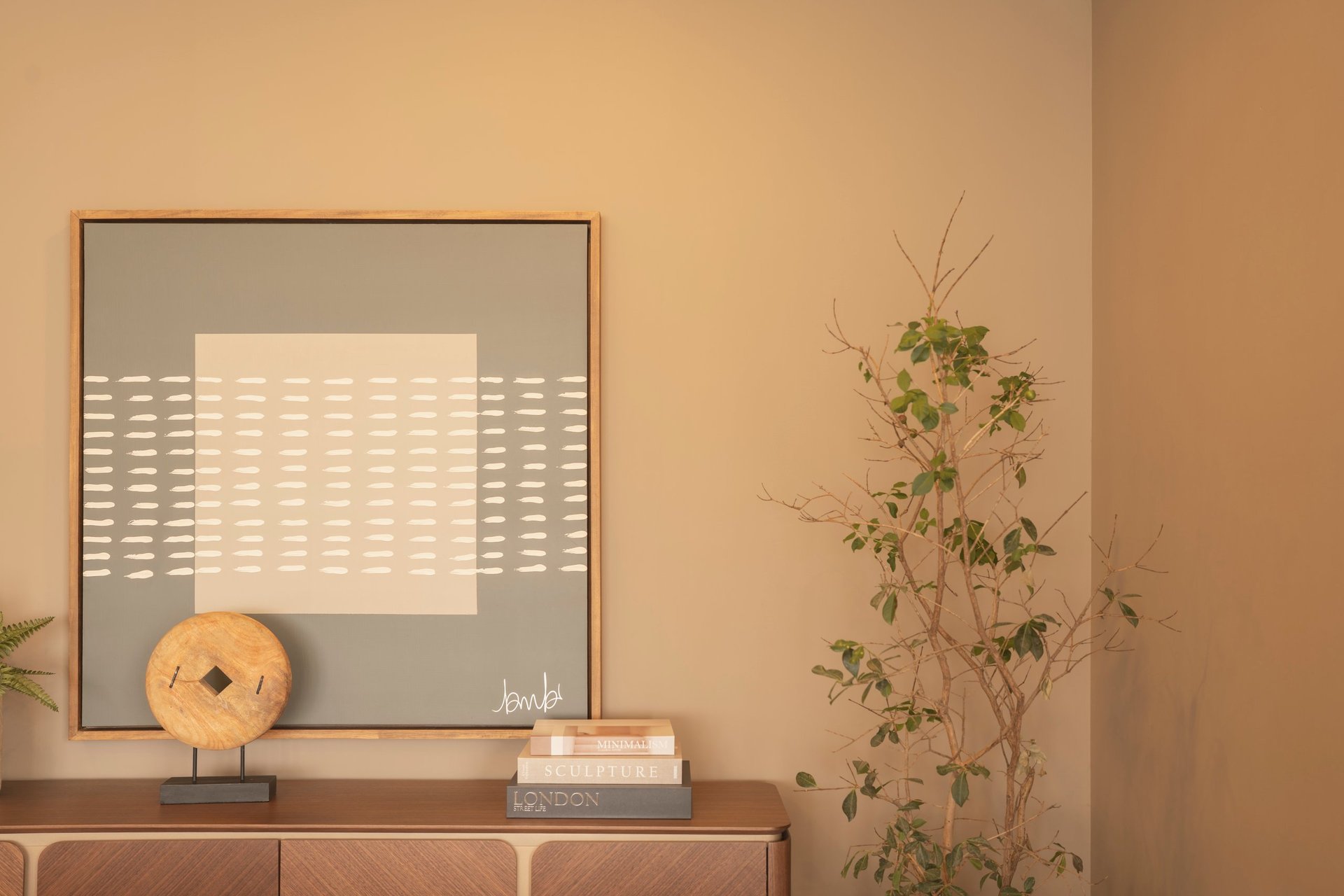 Minimalist room with beige walls, a wooden dresser displaying a woven basket sculpture, stacked books, and a climbing plant against a warm monochromatic background