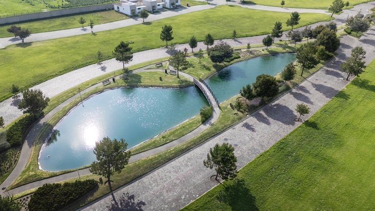 Aerial view of a modern landscape with a curved blue pond, walking paths, green lawn, and scattered trees in an urban park or residential development.