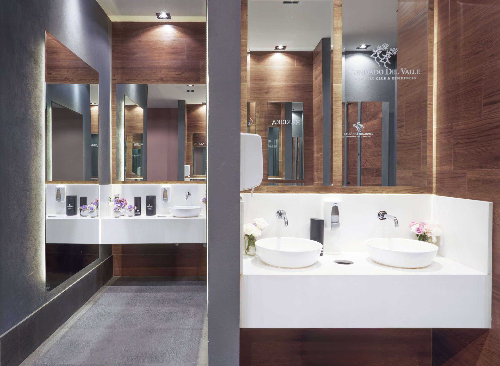Modern bathroom with dual white sinks, large mirrors, brown wood walls, and glass shower enclosure with ambient lighting