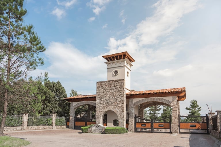 Stone and stucco entrance gate with a clock tower, wooden arches, and metal gates, surrounded by trees and landscaping under a partly cloudy sky
